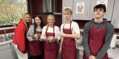 students wearing red aprons hold up a baking pan filled with brookie batter