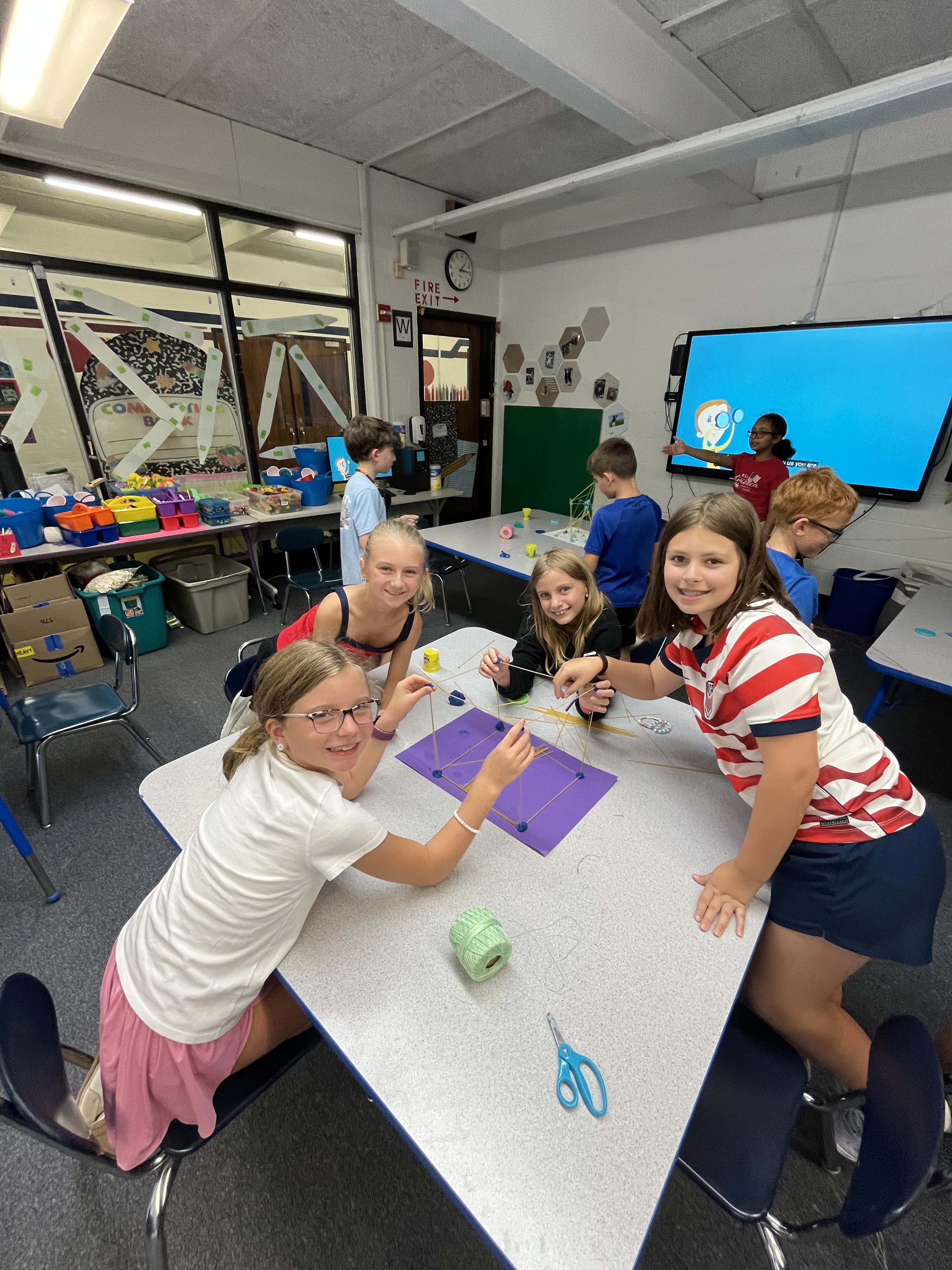 Students smile and stand around desk while working on their tower