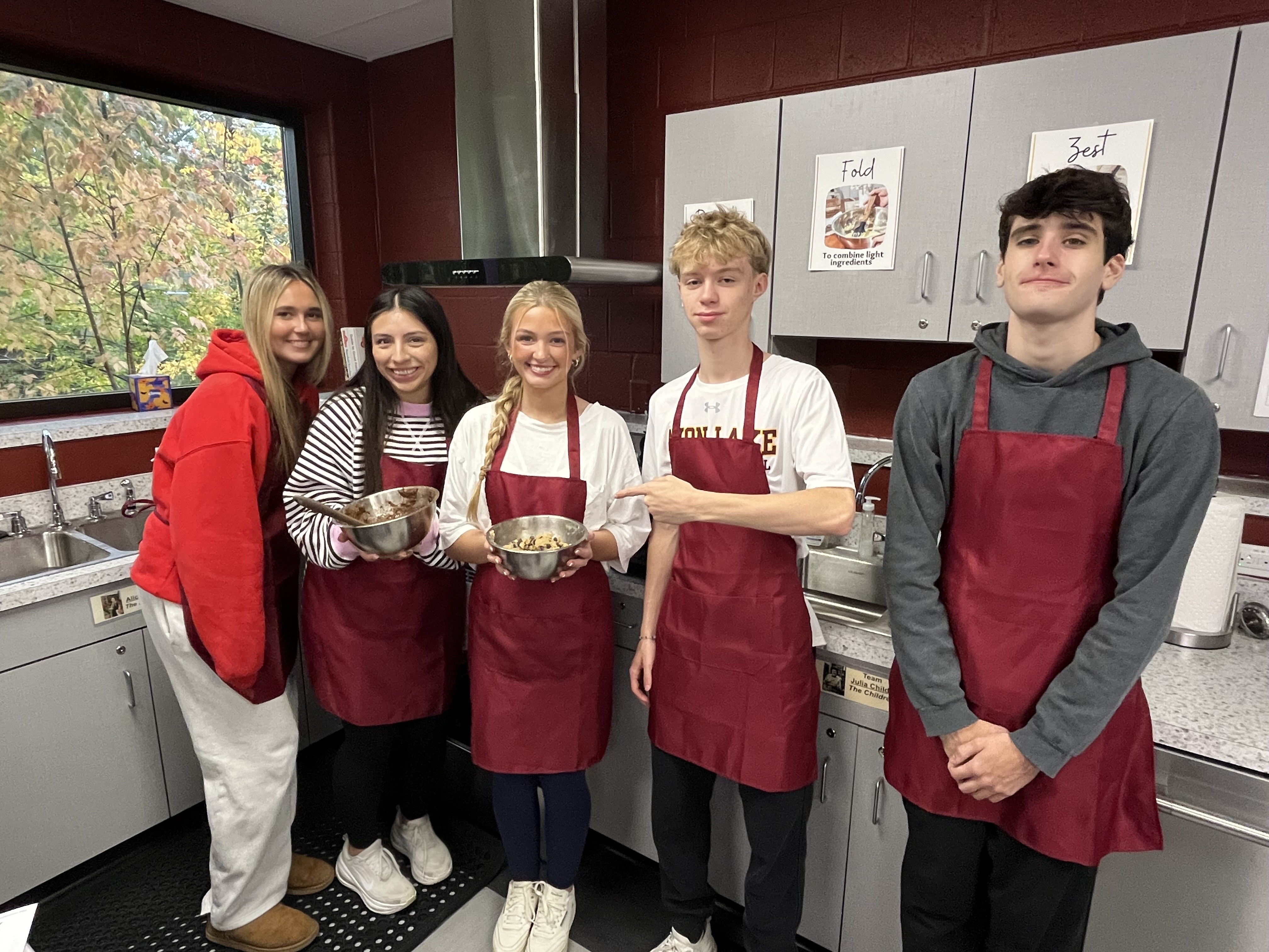 students wearing red aprons hold up baking pan filled with brookie mix